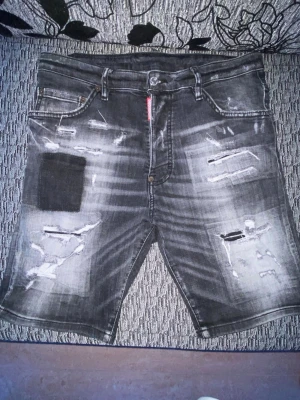 Svarta slitna jeansshorts DSO2 - Säljer ett par svarta jeansshorts från DSO2 med slitningar och patchdetaljer framtill. Shortsen har fem fickor, bälteshällor och en röd logga bak i midjan. Perfekta för dig som gillar en edgy streetstil.