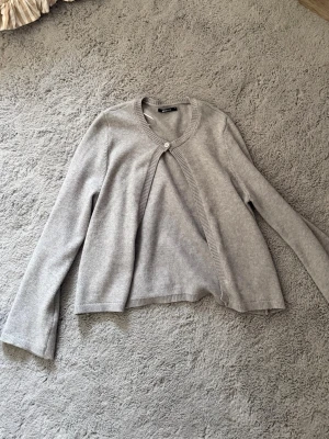 Ljusgrå kofta från Gina Tricot - Ljusgrå kofta från Gina Tricot med rund halsringning och en knapp framtill. Koftan har långa ärmar och är tillverkad i ett mjukt, tunt stickat material. Perfekt att slänga över en topp för en enkel och clean look.