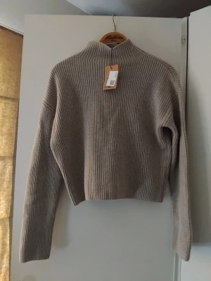 Beige ribbstickad tröja med hög krage - Säljer en beige ribbstickad tröja med hög krage och långa ärmar. Tröjan har en rak, något boxig passform och är tillverga kad i ett mjukt material som passar perfekt för kyligare dagar. Enkel och stilren design som är lätt att matcha.