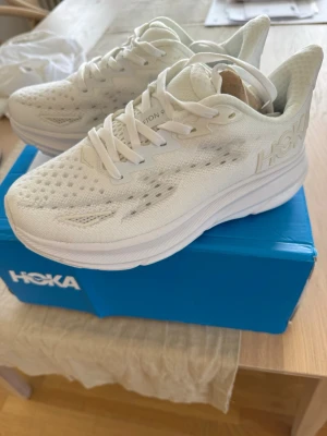Vita Hoka Clifton 9 sneakers - Säljer ett par vita Hoka Clifton 9 sneakers med ventilerande mesh-överdel och tjock, dämpad sula. Skorna har klassisk snörning och diskret Hoka-logga på sidan. Perfekta för dig som gillar stilrena och bekväma sneakers.