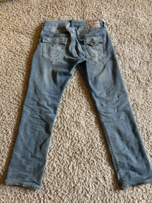 Ljusblå True Religion jeans - Snygga ljusblå jeans från True Religion med klassiska bakfickor och kontrastsömmar. Jeansen har raka ben och detaljerade ficklock med knapp. Perfekta för dig som gillar streetwear och vill sticka ut med ett ikoniskt märke.