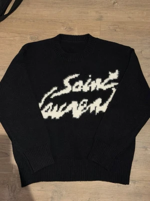 Svart Saint Laurent stickad tröja - Säljer en svart stickad tröja från Saint Laurent med vit grafisk logga framtill. Tröjan har rund halsringning, långa ärmar och breda ribbade muddar vid ärmslut och nederkant. Perfekt för dig som gillar streetstyle och exklusiva märken. Storlek M. Priset kan alltid diskuteras
