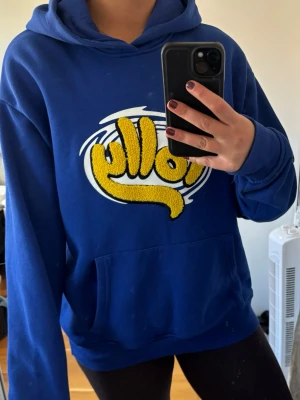 Blå Polly hoodie med gul logga - Säljer en blå hoodie med stor gul Polly-logga i frotté på bröstet. Hoodien har huva och en klassisk magficka framtill. Perfekt för dig som gillar färgglada och unika plagg. Materialet är mjukt och skönt, troligen bomullsblandning.