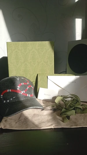  Gucci keps  - Snygg svart keps från Gucci med ikoniskt ormtryck i rött, vitt och svart på framsidan. Kepsen har klassisk böjd skärm och mesh på baksidan för extra ventilation. Justerbar rem baktill och diskret GG-mönster i tyget ger en exklusiv känsla.