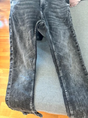 Svarta tvättade jeans från SMOG - Säljer ett par svarta tvättade jeans från SMOG med snyggt slitna detaljer och raka ben. Jeansen har klassisk femficksmodell och silverfärgad knapp med logga. Perfekta för en avslappnad och trendig look.