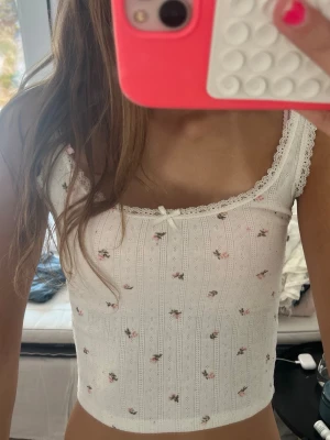 Vit blommig linnetopp med spets - Supersöt vit linnetopp med små blommor och spetsdetaljer längs kanterna. Toppen har en liten rosett framtill och är croppad i modellen. Perfekt för sommaren.