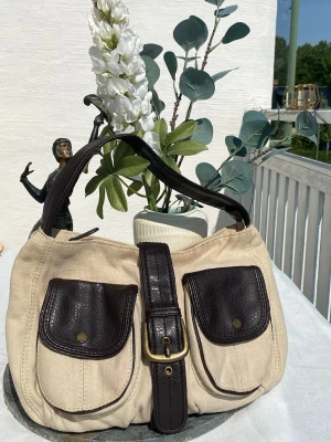 Beige axelremsväska med bruna detaljer💞☀️ - Snygg beige handväska i canvas med mörkbruna detaljer i läderimitation. Väskan har två framfickor med lock och tryckknappar samt ett brett axelband. Stängs med ett dekorativt spänne framtill. Perfekt för dig som gillar retrovibbar och praktiska fack. Se sita bild för skick.