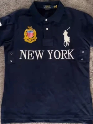 Marinblå piké från Polo Ralph Lauren. Chief Keef piké   - Snygg marinblå pikétröja från Polo Ralph Lauren med vita detaljer, stor logga och texten 'NEW YORK' på bröstet. Klassisk krage, knappar framtill och stort vitt nummer 5 på ryggen. Perfekt för en sportig och stilren look.