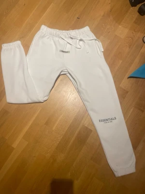 Vita mjukisbyxor från Essentials - Säljer ett par vita mjukisbyxor från Essentials med tryck på benet och dragsko i midjan. Byxorna har resår i midjan och vid bensluten för en skön passform. Perfekta för chill dagar eller när du vill ha en clean look.