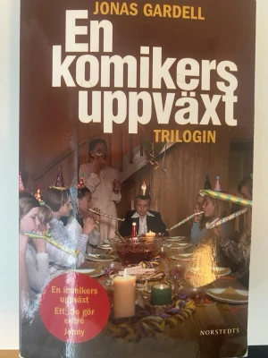 En komikers uppväxt Trilogin - En samlingsvolym av Jonas Gardells klassiska trilogi 'En komikers uppväxt'. Omslaget visar ett barnkalas med färgglada partyhattar och serpentiner. Boken är i pocketformat och utgiven av Norstedts.