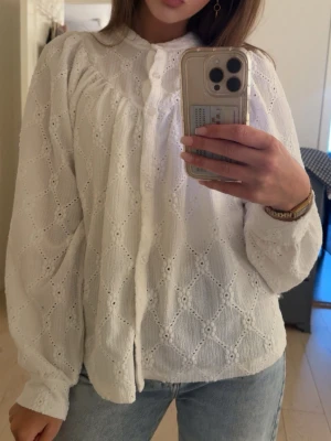 Vit broderad blus med puffärm - Söt vit blus med broderade detaljer och knappar framtill. Blusen har puffiga långa ärmar och rundad krage. Tillverkad i ett lätt och luftigt tyg med hålmönster som ger en romantisk känsla. Perfekt att styla med jeans eller kjol för en trendig look.