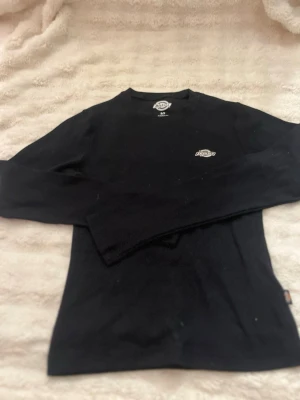 Svart långärmad tröja från Dickies - Svart långärmad tröja från Dickies med rund hals och liten vit logga på bröstet. Tröjan har en enkel och clean design, perfekt att matcha med jeans eller byxor. Tillverkad i mjukt bomullsmaterial för skön känsla.