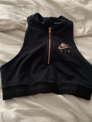 Svart croppad linnetopp från Nike Air känns som S! - Snygg svart croppad linnetopp från Nike Air med guldig dragkedja framtill och logga i guld på bröstet. Perfekt för en sportig look och passar till både jeans och träningsbyxor. Hög hals och ärmlös design ger en modern vibe.