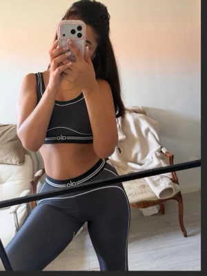 Svart träningsset från Alo - Snyggt svart träningsset från Alo med vita kontrastsömmar. Setet består av en sport-bh och höga tights, båda med elastisk midja och logga framtill. Perfekt för gymmet eller yoga. Materialet är stretchigt och följsamt för maximal komfort.