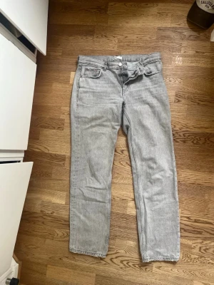 Grå raka jeans med rå kant - Säljer ett par ljusgrå jeans med rak passform och klassisk femficksdesign. Jeansen har rå kant nertill och stängs med knappgylf. Materialet är denim i bomull och färgen är tvättad grå, perfekt för en avslappnad look.