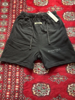 Svarta shorts Essentials Fear of God - Svarta shorts från Essentials Fear of God med elastisk midja och långa snören framtill. Shortsen har tryckt logga på vänster ben och en bakficka. Tillverkade i mjukt bomullsmaterial för en avslappnad look.