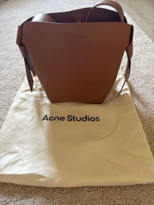 Acne studios musibi mini - Snygg brun handväska från Acne Studios i stilren bucket-modell. Väskan är gjord i slätt läder och har bred axelrem samt dekorativa knutar på sidorna. Perfekt för dig som gillar minimalistisk design och vill ha en trendig accessoar.