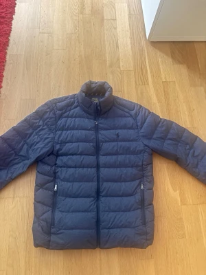 Mörkblå dunjacka med dragkedja - Snygg mörkblå dunjacka med ståkrage och två dragkedjeförsedda fickor framtill. Jackan har quiltad design med horisontella sömmar och diskret logga på bröstet. Perfekt för kalla vinterdagar och enkel att matcha med olika outfits.