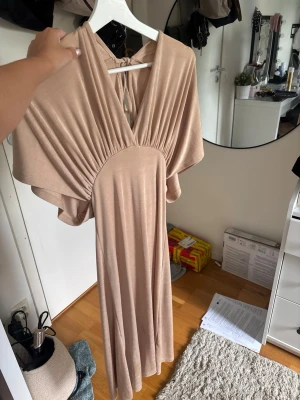 Beige långklänning med v-ringning - Säljer en elegant beige långklänning med djup v-ringning och draperad överdel. Klänningen har vida, fladdriga ärmar och knytning i nacken. Materialet är mjukt och faller snyggt längs kroppen, perfekt för en trendig look.