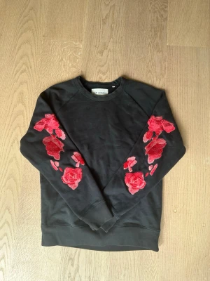 Svart sweatshirt med röda rosor - Svart sweatshirt med broderade röda rosor på båda ärmarna. Tröjan är från our legacy och storleken är 46 och passar M/S