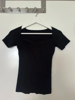 Svart ribbad topp med fyrkantig ringning - Snygg svart ribbad topp med korta ärmar och fyrkantig ringning. Toppen har en tight passform och är tillverkad i stretchigt material som sitter skönt på kroppen. Perfekt att styla med jeans eller kjol för en trendig look.