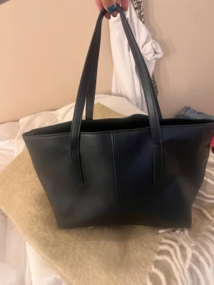 Svart handväska  - Säljer en svart handväska i skinnimitation med två långa handtag. Väskan har en enkel och stilren design. Perfekt storlek för att få plats med det viktigaste och passar till många olika outfits💗