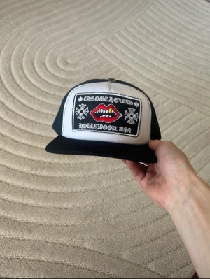 Chrome Hearts truckerkeps svart/vit - Säljer en svart och vit truckerkeps från Chrome Hearts med mesh baktill och justerbar snapback. Framtill finns en stor patch med röda läppar, vita tänder och texten 'Chrome Hearts Hollywood, USA' samt blommönster. Perfekt statement piece för streetwear-stilen.
