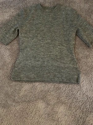 Grå stickad tröja med kort ärm - Mysig grå stickad tröja med korta ärmar och rund halsringning. Tröjan har bred ribbstickad kant nedtill och vid ärmsluten. Perfekt för lager på lager och enkel att matcha med olika stilar.