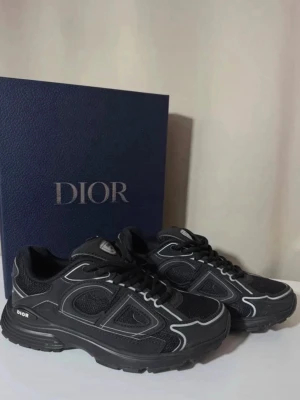 Dior B30 svarta - Snygga svarta Dior sneakers med vita kontrastsömmar och robust sula. Skorna har en sportig design med mesh och läderpaneler samt logga på sidan och plösen. Perfekta för dig som vill ha en exklusiv och trendig look.