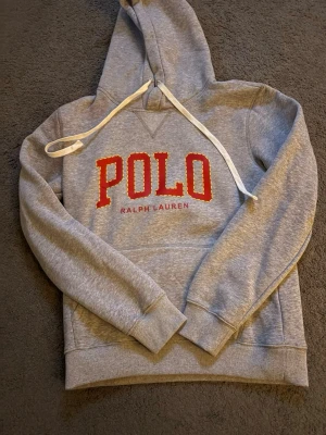 Grå hoodie från Polo Ralph Lauren - Snygg grå hoodie från Polo Ralph Lauren med stor röd POLO-logga och text på bröstet. Klassisk huva med vita dragsnören och magficka framtill. Tillverkad i mjukt sweatshirtmaterial, perfekt för en avslappnad stil.