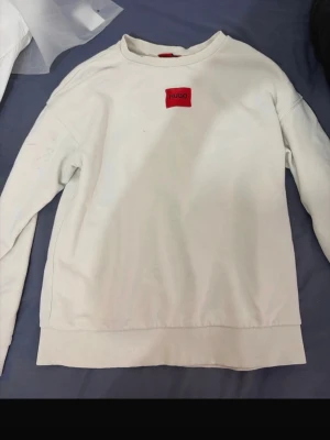 Vit sweatshirt från Hugo - Säljer en stilren vit sweatshirt från Hugo med röd patch och logga på bröstet. Tröjan har rund halsringning, ribbade muddar och är tillverkad i mjukt bomullsmaterial. Perfekt för en clean och enkel look.