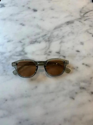 S&T 003 Green solglasögon  - Snygga solglasögon med transparent grågrön båge och runda, bruna glas. Klassisk och trendig form som passar till många stilar. Bågarna är i plast och glasen har en varm ton som ger en cool look.