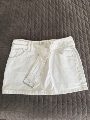 Vit kort jeanskjol med bälte - Snygg vit kort jeanskjol med tillhörande bälte och silverfärgat spänne. Kjolen har klassiska fickor fram och bak samt hällor i midjan. Perfekt för dig som vill ha en clean och fräsch look till sommaren.