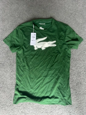 Grön Lacoste Sport t-shirt - Snygg grön t-shirt från Lacoste Sport med stor vit krokodil-logga på bröstet. Klassisk rund hals och korta ärmar. Regular fit för en avslappnad look. Perfekt för dig som gillar sportig och stilren design.