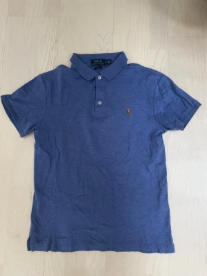 Blå pikétröja från Polo Ralph Lauren - Säljer en blå pikétröja från Polo Ralph Lauren i slim fit-modell. Tröjan har korta ärmar, klassisk krage och knappar framtill. Ikoniska broderade loggan på bröstet. Perfekt för en stilren och avslappnad look. Storlek M