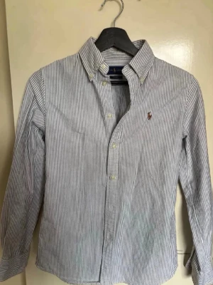 Randig skjorta från Ralph Lauren - Klassisk skjorta från Ralph Lauren med smala blå och vita ränder. Skjortan har button-down krage, långa ärmar och den ikoniska broderade loggan på bröstet. Tillverkad i bomull för en skön och stilren look.