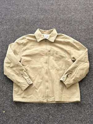 Beige/grön overshirt från Bershka - Stilren beige overshirt från Bershka i bomullstyg. Modellen har dragkedja framtill, en bröstficka och klassisk krage. Perfekt lager på lager-plagg med avslappnad passform och knappdetaljer vid ärmsluten.