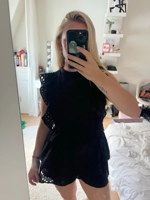 Svart broderad jumpsuit med volanger - Supersnygg svart jumpsuit med broderade detaljer och volanger längs axlarna. Modellen har markerad midja med knytskärp och korta ben. Perfekt för dig som vill ha en trendig och bekväm look. Materialet är lätt och luftigt.