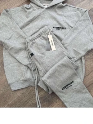 Grått Essentials set med hoodie & byxor - Snyggt grått set från Essentials med hoodie och matchande mjukisbyxor. Båda plaggen har svart Essentials-logga tryckt på framsidan. Hoodien har huva och byxorna har dragsko i midjan för skön passform. Perfekt för en avslappnad och trendig look.