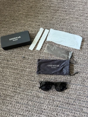 Svarta solglasögon från ONEWear - Snygga svarta solglasögon från ONEWear med klassisk wayfarer-form. Glasögonen har svarta bågar och mörka linser. Medföljer originalfodral, två tygpåsar och putsdukar. Perfekta för att höja din stil i solen.