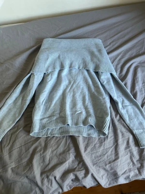 Ljusblå off shoulder stickad tröja - Trendig ljusblå stickad tröja med bred ribbad off shoulder-krage och långa ärmar. Tröjan har ribbade muddar vid ärmslut och nederkant. Perfekt för dig som vill ha en stilren och bekväm look.