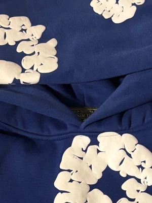 Blå hoodie Denim tears  - Säljer en blå hoodie med stora vita blommönster tryckta över hela plagget. Hoodien har en klassisk huva och är tillverkad i mjuk bomull. Perfekt för dig som vill sticka ut med en unik och färgglad stil.