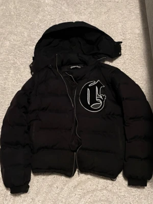 Svart pufferjacka från Corvidae - Snygg svart pufferjacka från Corvidae med stor vit broderad logga på bröstet. Jackan har avtagbar huva och dragkedja framtill. Perfekt för kalla vinterdagar och ger en streetwear vibe till din outfit.