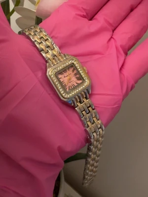 Guldig och silverfärgad klocka med rosa urtavla - Snygg klocka med fyrkantig urtavla i rosa och romerska siffror. Boetten är dekorerad med gnistrande stenar och armbandet är i både guld- och silverfärg. Perfekt accessoar för att lyfta din stil.