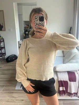 Beige stickad tröja med puffärm - Säljer en beige stickad tröja med rund halsringning och puffiga ärmar. Tröjan har ribbade muddar vid ärmslut och nederkant, vilket ger en snygg och avslappnad look. Perfekt att matcha med jeans eller kjol för en trendig outfit.