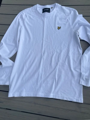 Vit långärmad tröja från Lyle & Scott - Vit långärmad topp från Lyle & Scott med rund halsringning och gul broderad logga på bröstet. Tillverkad i mjuk bomull för en clean och enkel stil. Perfekt för dig som gillar klassiska och stilrena plagg. Den är inte använd, hör av dig vid frågor. Billigare pris vid snabb affär!