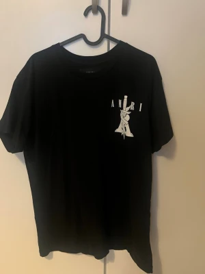 Svart AMIRI t-shirt med tryck - Säljer en svart t-shirt från AMIRI med ett stort vitt tryck av en hand som håller ett svärd och texten AMIRI både fram och bak. Klassisk passform och rund hals. Perfekt för dig som gillar streetwear och vill sticka ut. Den ligger bara hemma och använder aldrig den så vill bli av med den. Ny kostar över 3500kr men pris kan diskuteras 