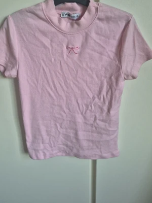 Ljusrosa t-shirt med rosett - Supersöt ljusrosa t-shirt med liten broderad rosett framtill. T-shirten har korta ärmar. Perfekt för dig som gillar gulliga detaljer och en mjuk, bekväm stil.