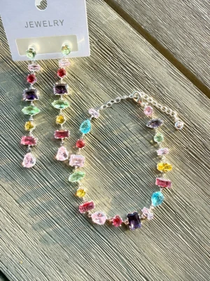 Färgglatt armband och örhängen med gnistrande stenar - Säljer ett armband  och örhängen med gnistrande stenar i flera färger som blått, rosa, gult, grönt, rött och lila. Armbandet har silverfärgad kedja och justerbar längd. Örhänget längd 7 cm. Stenarna är slipade i olika former för en unik och lekfull look.