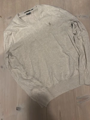 Ljusgrå v-ringad tröja - Ljusgrå v-ringad tröja med lång ärm och diskret broderad logga på bröstet. Tröjan har ribbade muddar vid ärmslut och nederkant. Perfekt för lager-på-lager och enkel att matcha med olika outfits.